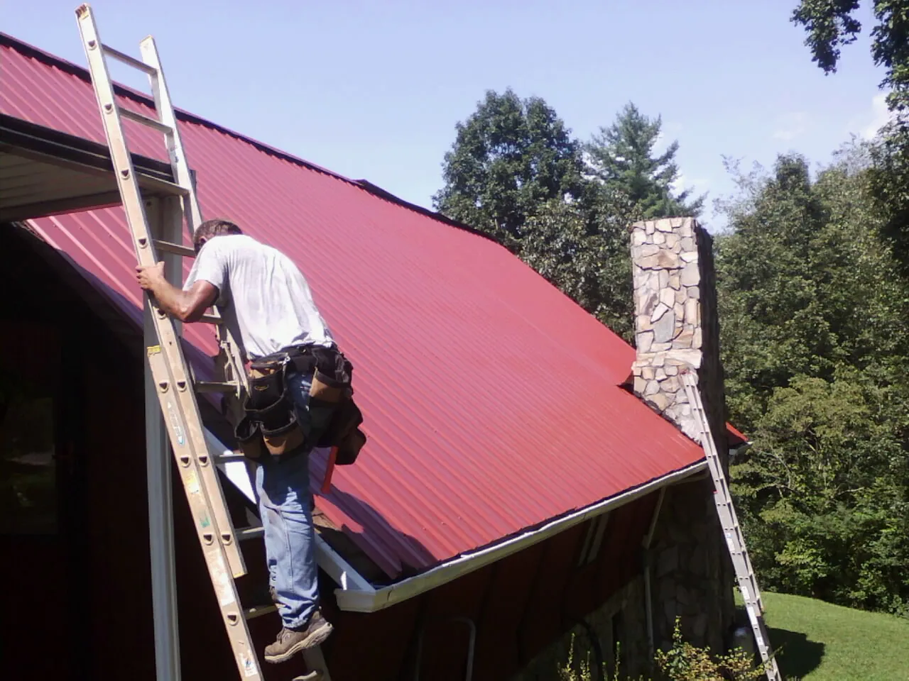 Roofer on a ladder beside a red metal roof and stone chimney — Soffit & Fascia Repair by Roofing Brothers in Harveyville